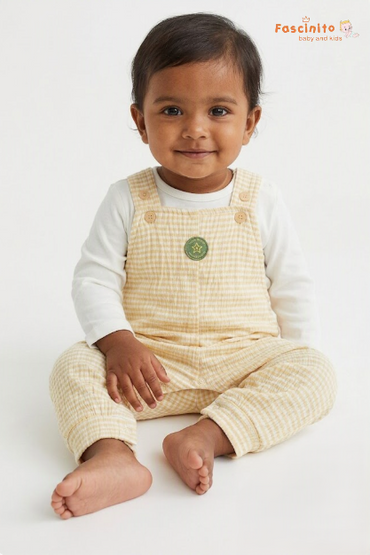 A baby sitting on a white studio background wearing a cream and yellow gingham overall romper over a white long-sleeve shirt. The brand logo 'Foscinito baby and kids' is visible in the top right corner.